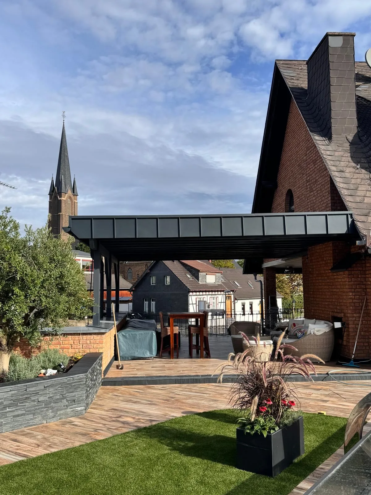 Neues Terrassendach mit Anthrazit-Stehfalzdeckung vor einem Backsteinhaus mit Schieferdach